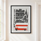 A framed Edinburgh cityscape art print hanging on a white wall beside a window. The poster shows black architectural silhouettes and the word 'EDINBURGH' vertically, with a red and white double-decker bus at the bottom.