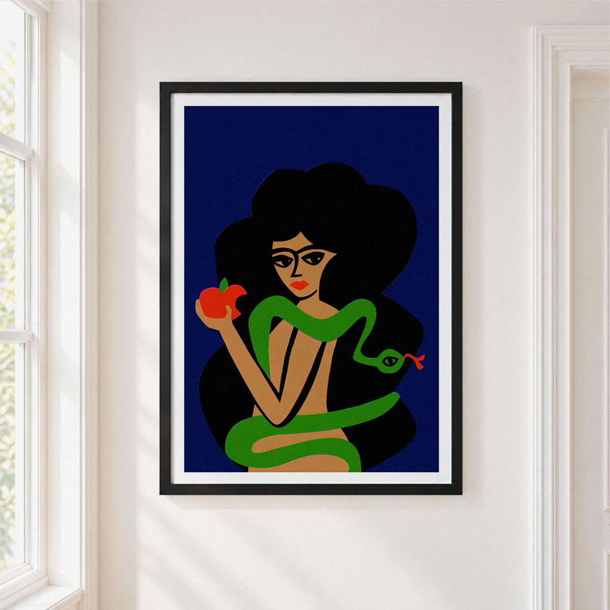A framed art print featuring a stylized illustration of a woman with glasses, black hair, and a green snake wrapped around her, holding a red apple, displayed on a white wall next to a window.