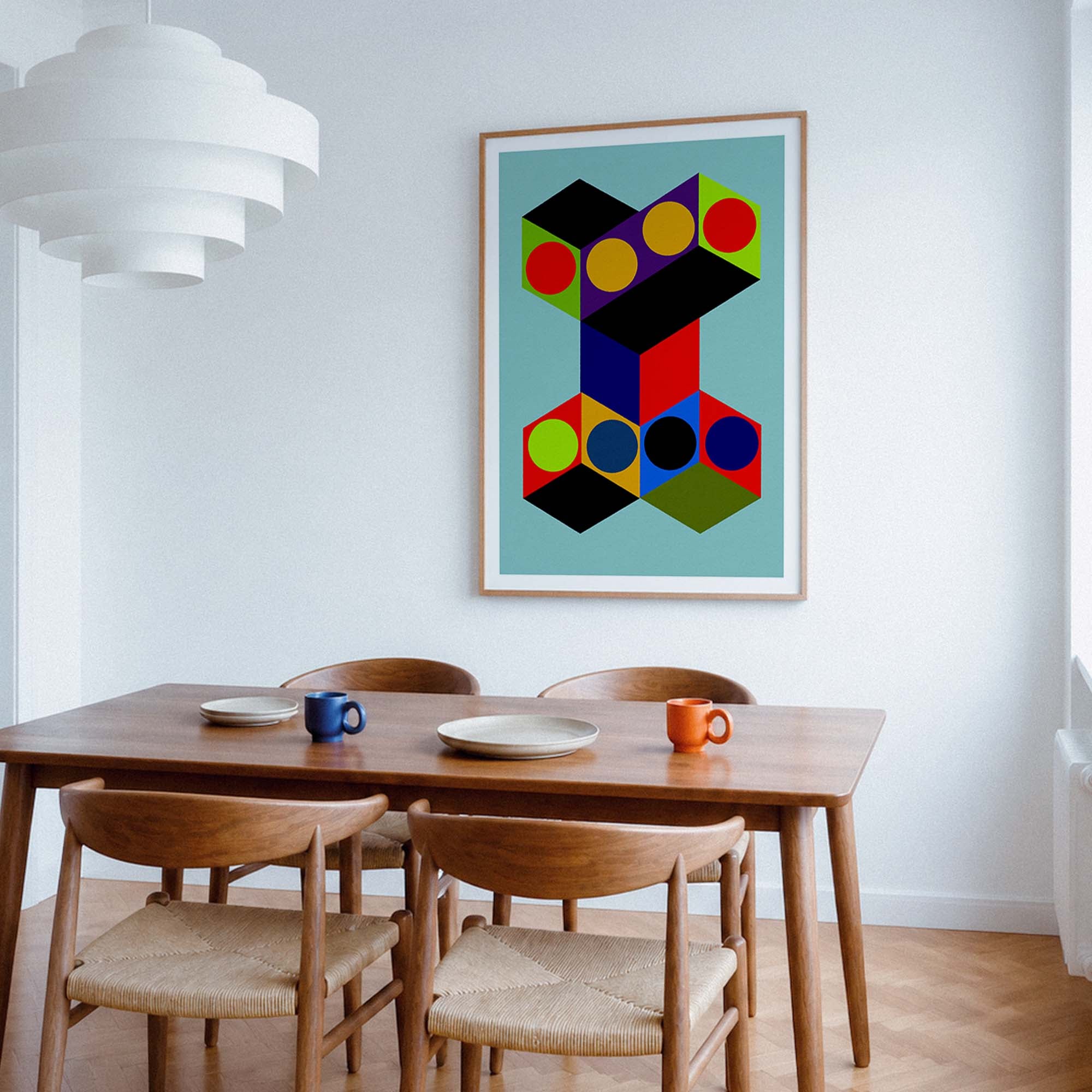 A framed abstract geometric art print hangs on a white wall above a wooden dining table with four woven-back chairs. The print features colorful 3D cube shapes on a light blue background, and the room is styled with a white pendant light, mugs, and plates.