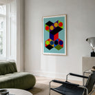 A framed abstract geometric art print with colorful 3D block shapes hangs on a light gray wall in a modern living room setting with a green sofa and black chair.