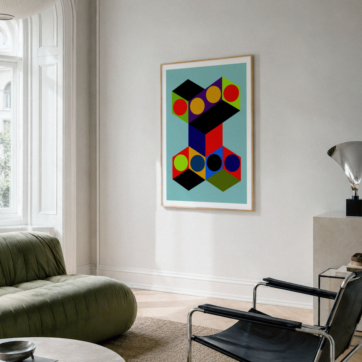 A framed abstract geometric art print with colorful 3D block shapes hangs on a light gray wall in a modern living room setting with a green sofa and black chair.