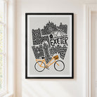 Framed art print featuring a black and white illustrated cityscape with the word 'EXETER' and an orange bicycle, displayed on a light beige wall in a real interior setting.