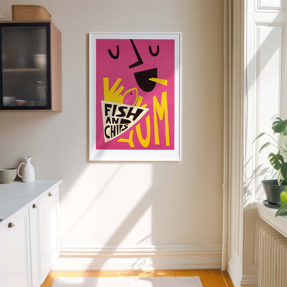 A framed art print featuring a colorful abstract fish and chips design on a pink background, hanging on a beige wall in a bright kitchen setting.