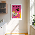 A framed art print featuring a colorful abstract fish and chips design on a pink background, hanging on a beige wall in a bright kitchen setting.