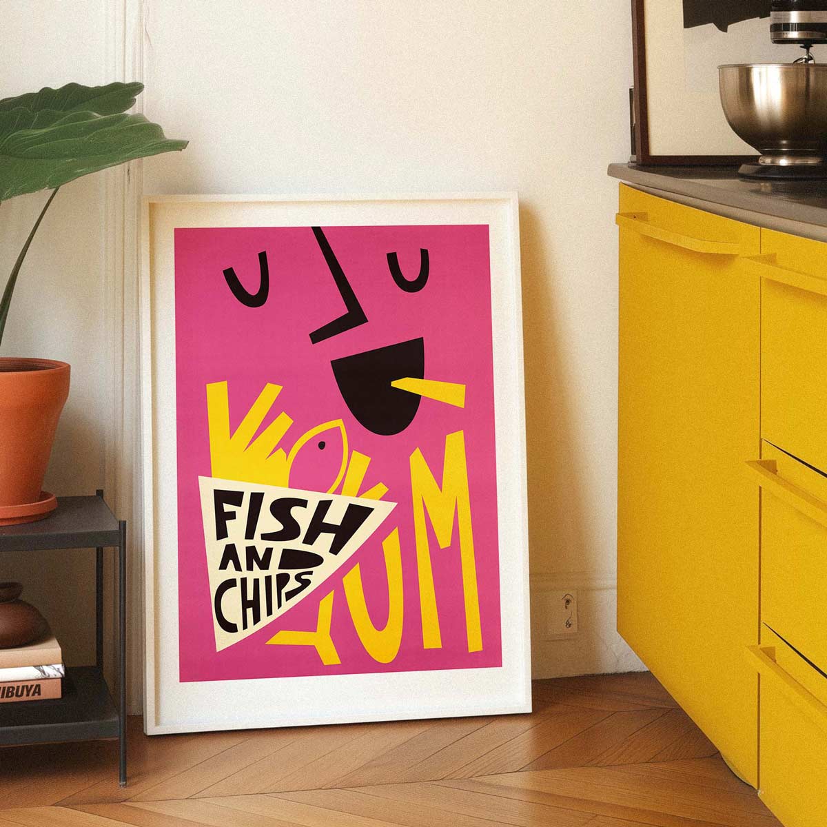 A framed art print with a colorful abstract design leaning against a wall in a kitchen setting. The print features a bright pink background with bold yellow, black, and white graphic elements and the text 'FISH AND CHIPS'.