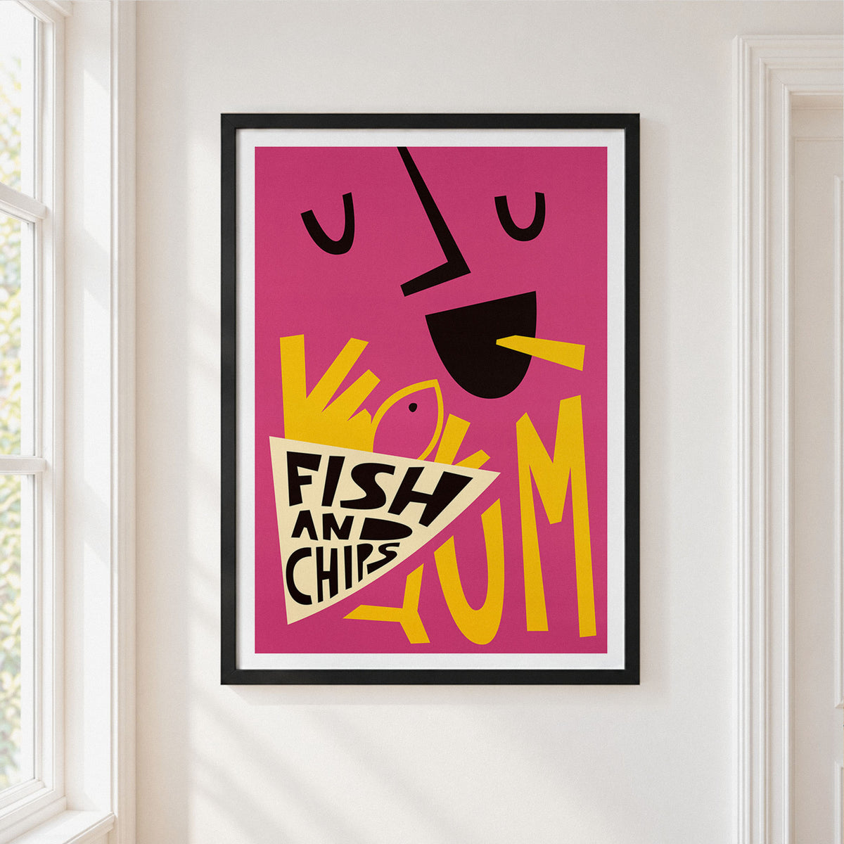A framed art print featuring a bold, retro-style illustration of a face eating chips with the words 'FISH AND CHIPS' on a pink background, hung on a white wall next to a window.