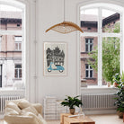 A framed art print featuring a light blue scooter and the word 'FLORENCE' in a black-and-white architectural illustration, hanging on a white wall in a bright, modern living room with large windows and plants.