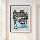 A framed art print featuring a stylized black-and-white illustration of Florence architecture with a light blue scooter, hanging on a cream-colored wall next to a window.