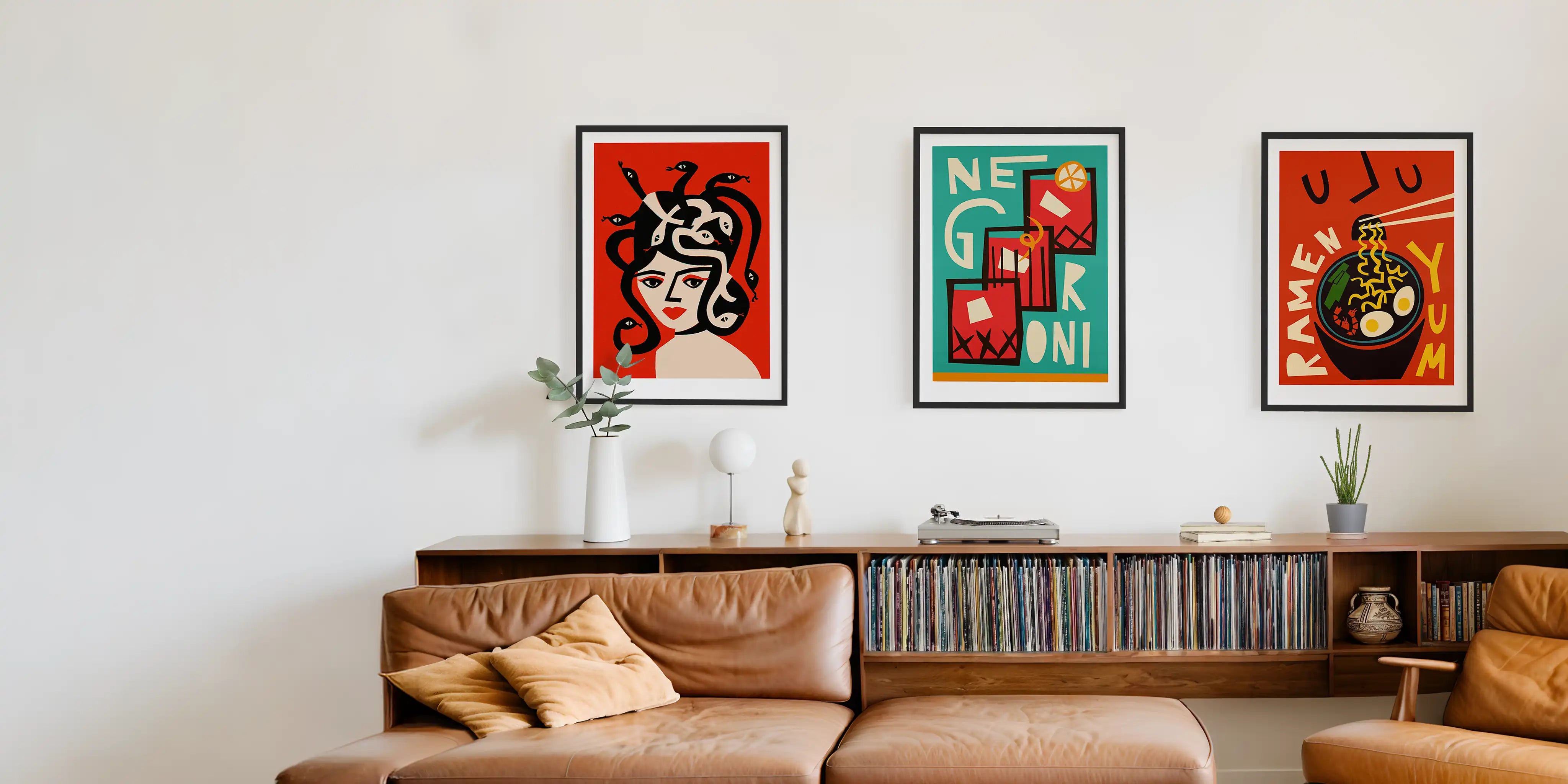 Three framed Fox & Velvet art prints with bold retro graphic designs are displayed on a white wall above a brown leather sofa in a modern living room setting. Medusa art print, Negroni art print and ramen art print.