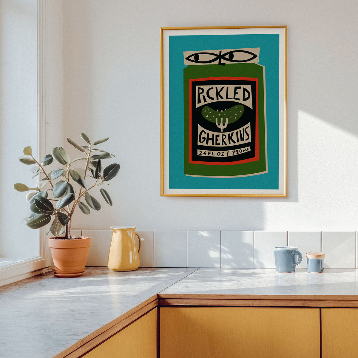 A framed art print featuring a retro-style green can graphic with the words 'PICKLED GHERKINS' and '22 FL OZ 710ML' hangs on a white kitchen wall above a countertop with a potted plant and ceramic pitchers.