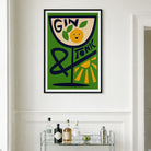 A framed vintage-style wall art print featuring a green background, a tilted glass with the words “GIN TONIC,” and an orange illustration, hanging on a white wall above a small bar cart with bottles and glasses.