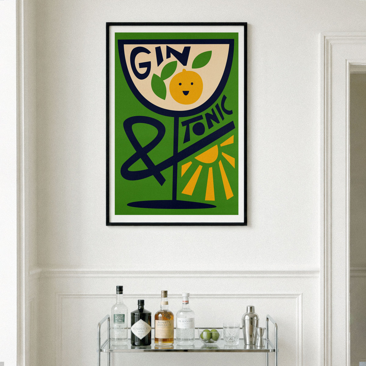 A framed vintage-style wall art print featuring a green background, a tilted glass with the words “GIN TONIC,” and an orange illustration, hanging on a white wall above a small bar cart with bottles and glasses.