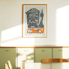 A framed art print featuring a stylized Glasgow skyline with a Orange train and black architectural details on a light gray background, hung on a cream-colored wall next to a window above a sideboard