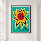 A framed art print featuring a stylized yellow sun with a red face on a teal background and the typography lettering 'GO FOR A WALK' at the bottom, hung on a white wall next to a window.