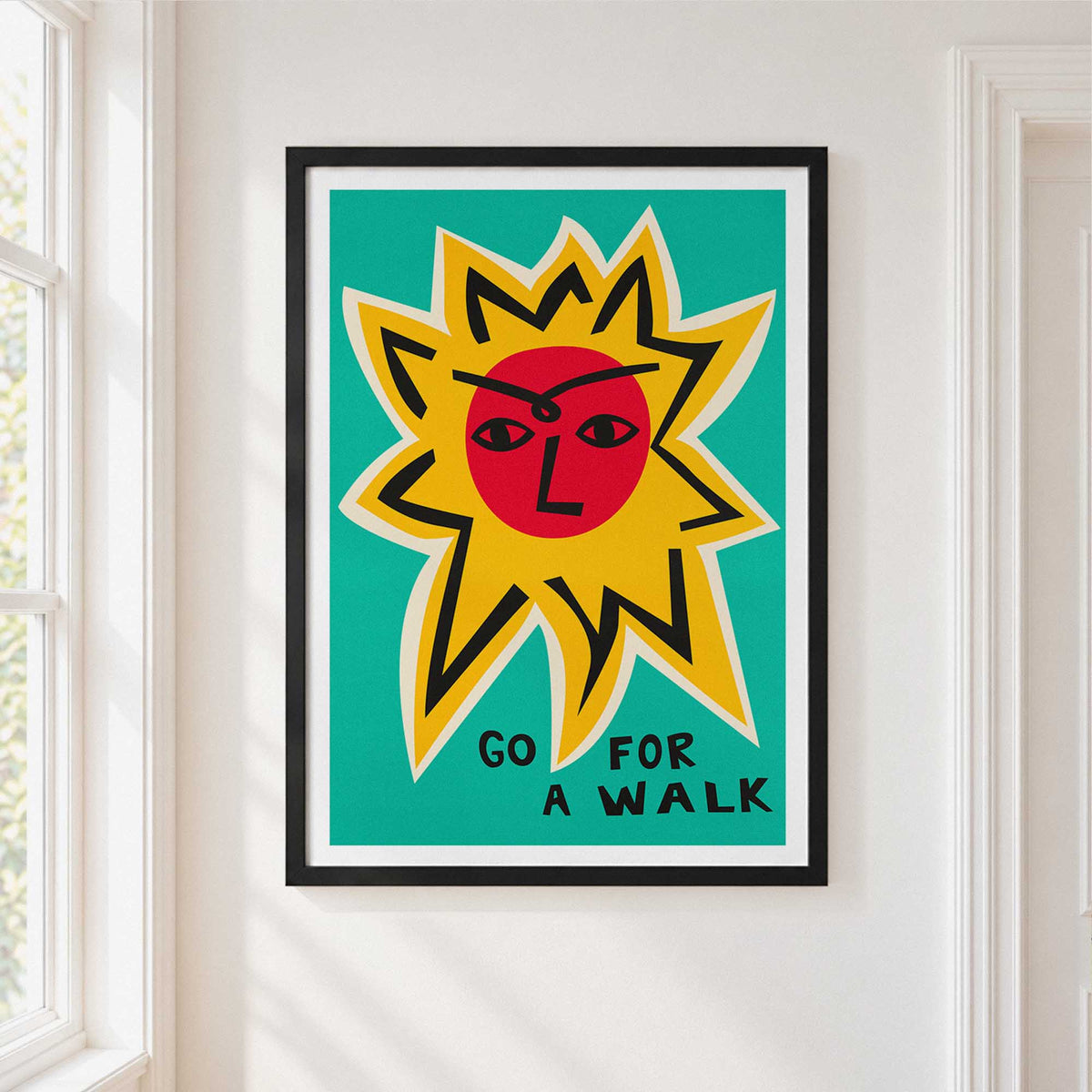A framed art print featuring a stylized yellow sun with a red face on a teal background and the typography lettering 'GO FOR A WALK' at the bottom, hung on a white wall next to a window.
