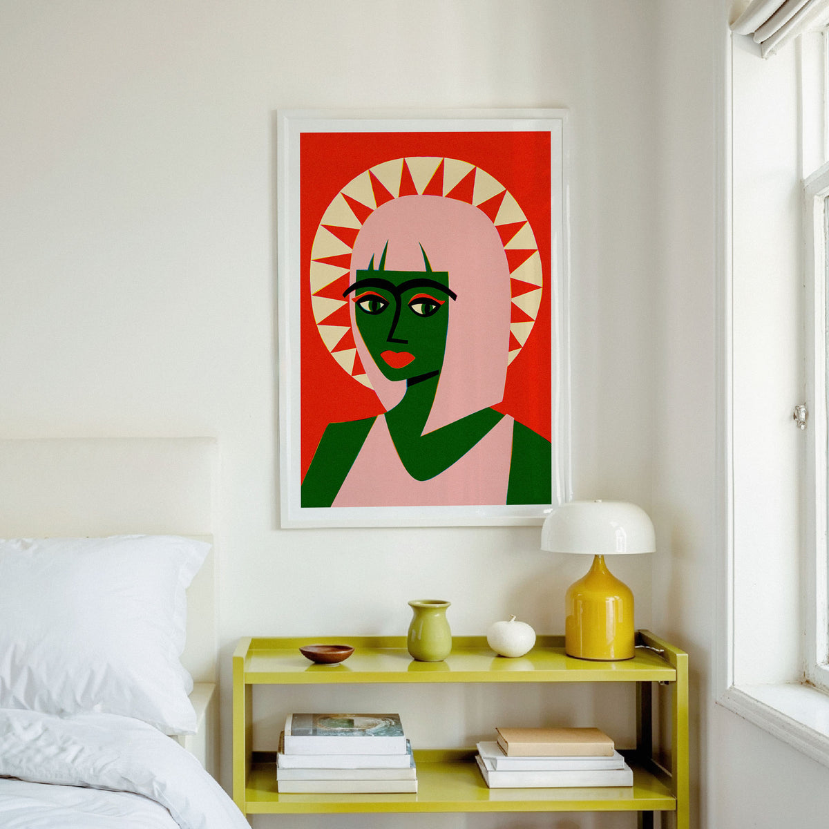 A framed art print featuring a stylized portrait of a woman with green skin, pink hair, and a red background, hanging on a white wall above a yellow bedside table in a bedroom setting.