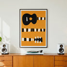 A framed abstract art print featuring a black guitar silhouette and striped shapes on a mustard yellow background, displayed on a wooden sideboard in a modern living room setting with speakers and a turntable.