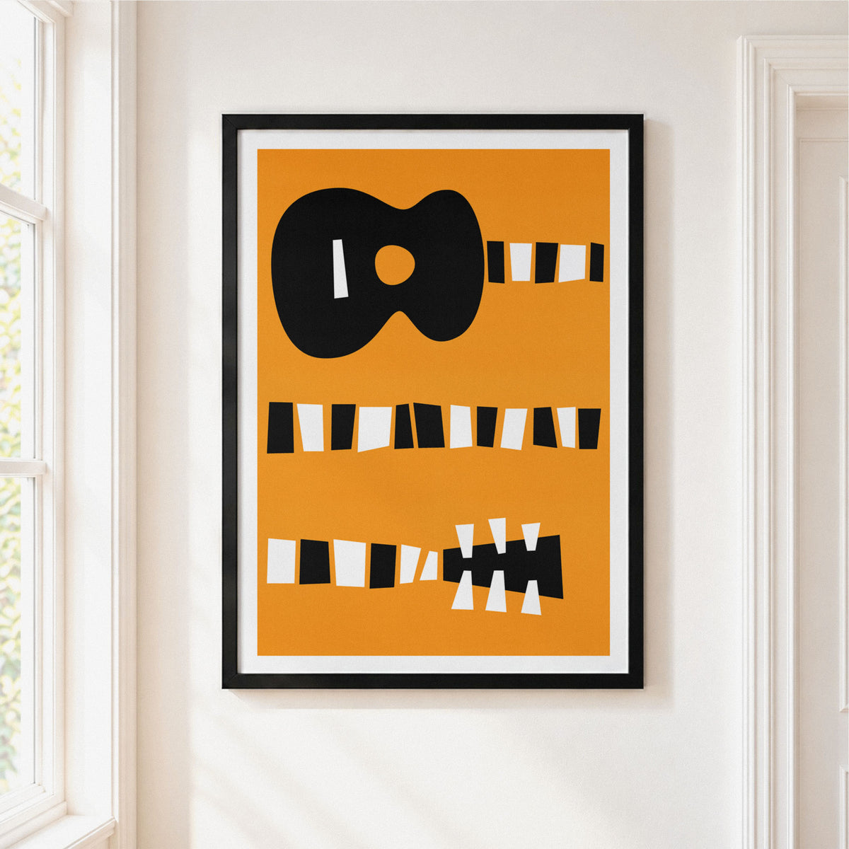 Framed minimalist art print featuring a stylized black guitar on a warm orange background, hung on a white wall next to a window.