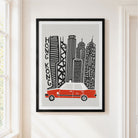 A framed art print featuring a stylized black and white illustration of Hong Kong skyscrapers with a red taxi at the bottom, hanging on a light beige wall next to a window.