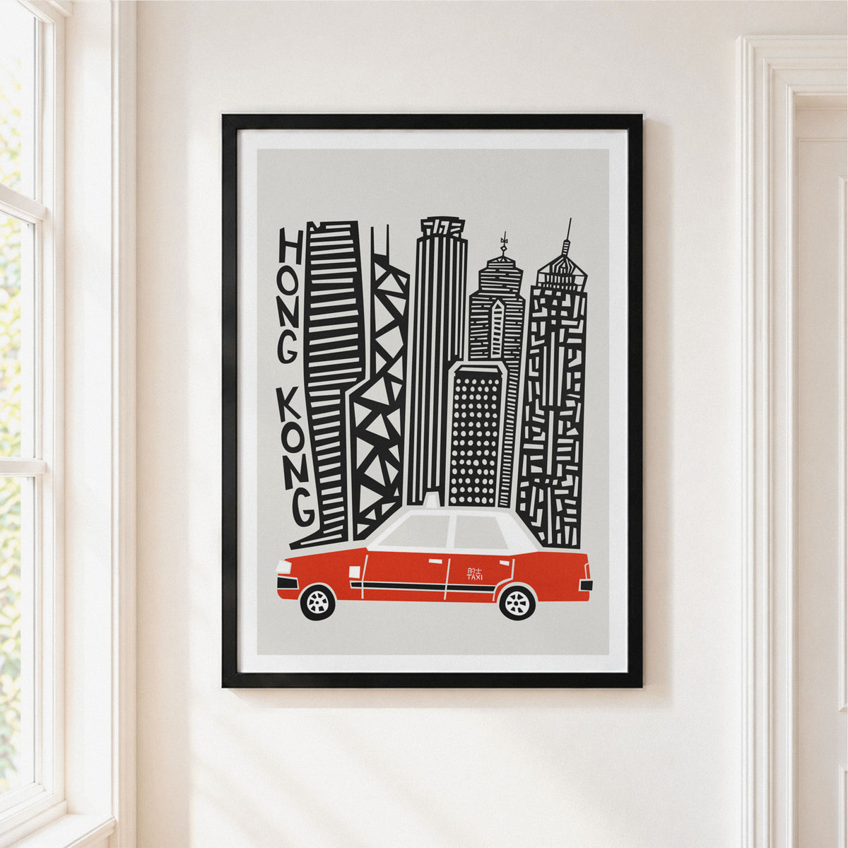 A framed art print featuring a stylized black and white illustration of Hong Kong skyscrapers with a red taxi at the bottom, hanging on a light beige wall next to a window.