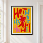A framed chilli's art print featuring a bold, retro-style graphic with the word 'HOT' repeated in red, pink, and blue on a yellow background, with a large red chili pepper in the center. The print is framed in black with a white mat, hanging on a white wall next to a window.