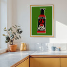 A framed art print featuring a retro hot sauce bottle on a green background, hanging on a white kitchen wall above a wooden counter with a potted plant and mugs.