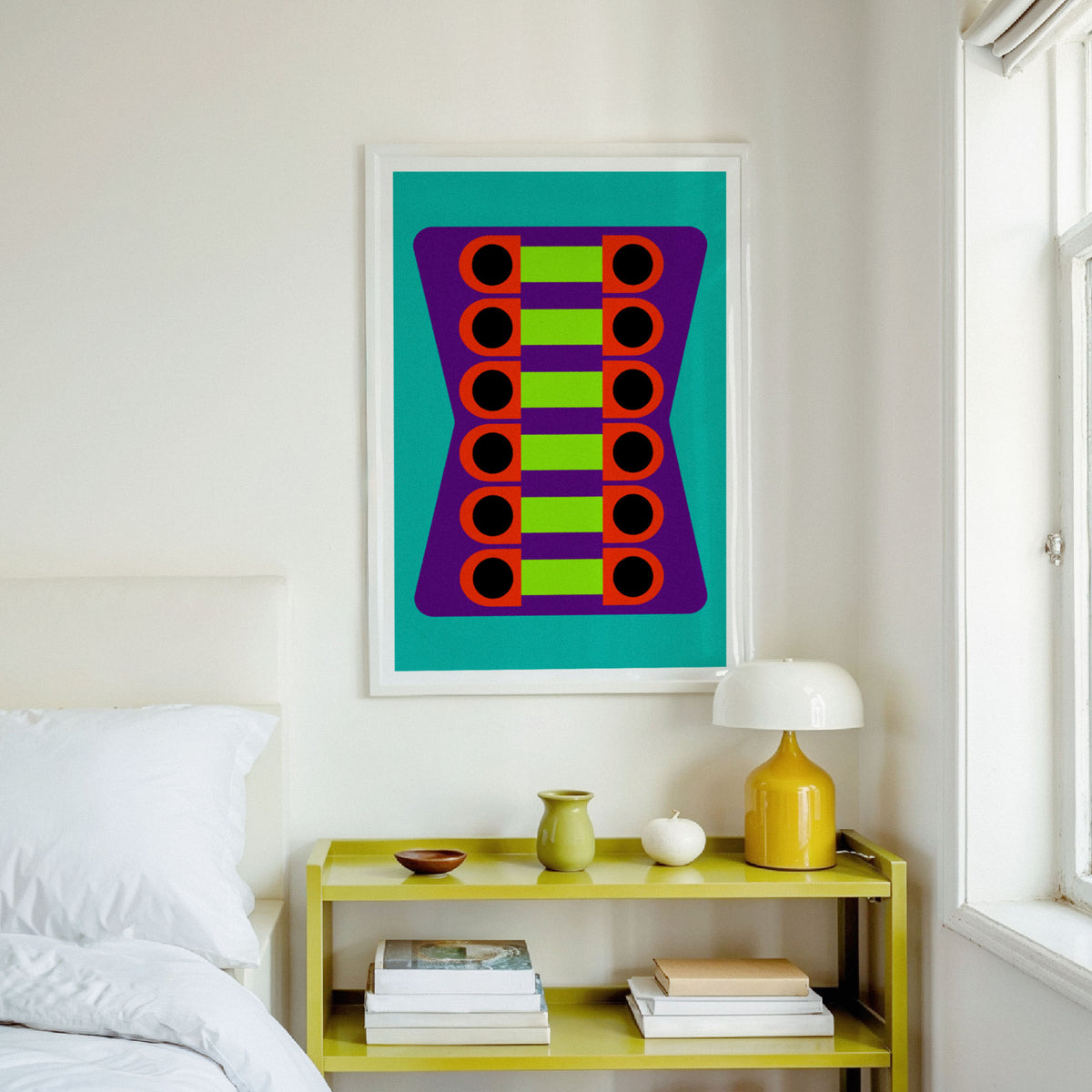A framed abstract art print featuring colorful geometric circles on a teal background, hanging on a white wall above a yellow shelf in a bright bedroom setting.