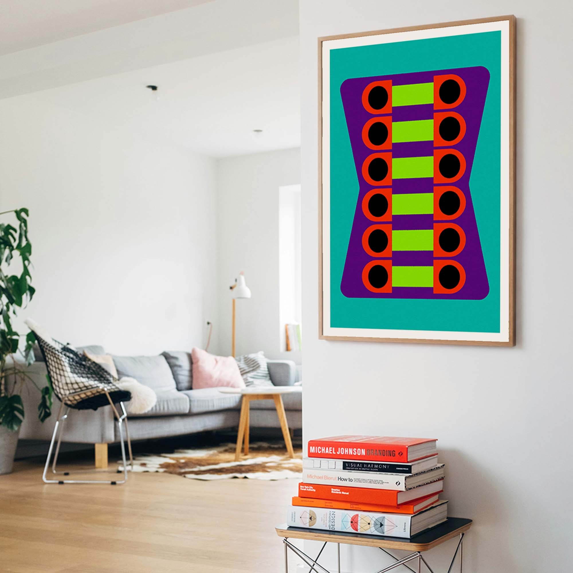 A framed abstract art print featuring a colorful geometric pattern of green, purple, orange, and black shapes on a teal background, displayed on a white wall in a modern living room setting.
