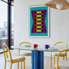 A framed abstract art print featuring a colorful geometric pattern hangs on a white wall in a modern dining room setting with a glass-top table, yellow chairs, and a hanging woven pendant light.