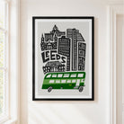 A framed art print featuring a stylized black and white city skyline with the word 'LEEDS' and a green double-decker bus, hanging on a light beige wall next to a window.
