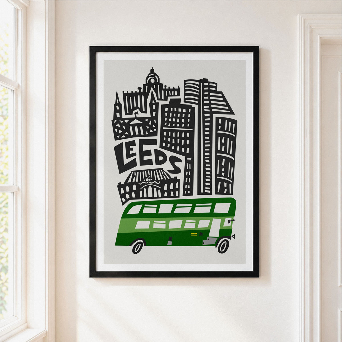 A framed art print featuring a stylized black and white city skyline with the word 'LEEDS' and a green double-decker bus, hanging on a light beige wall next to a window.