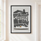 A framed Liverpool cityscape art print with black line art on a light gray background, hanging on a cream-colored wall next to a window.