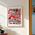 A framed art print featuring a stylized red and black illustration of London landmarks on a white background, hanging on a light beige wall in a sunlit room.