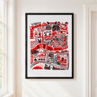 A framed art print featuring a red and black hand-drawn London cityscape with iconic landmarks, hung on a white wall next to a window.