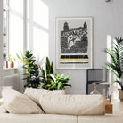 A framed Los Angeles cityscape art print with black and white illustration and a yellow stripe, displayed on a light gray wall above a beige sofa in a sunlit living room with potted plants.