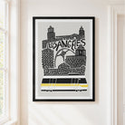 A framed Los Angeles cityscape art print hangs on a white wall next to a window. The print features black silhouettes of the skyline and a train on a light gray background, enclosed in a black frame with a white mat.