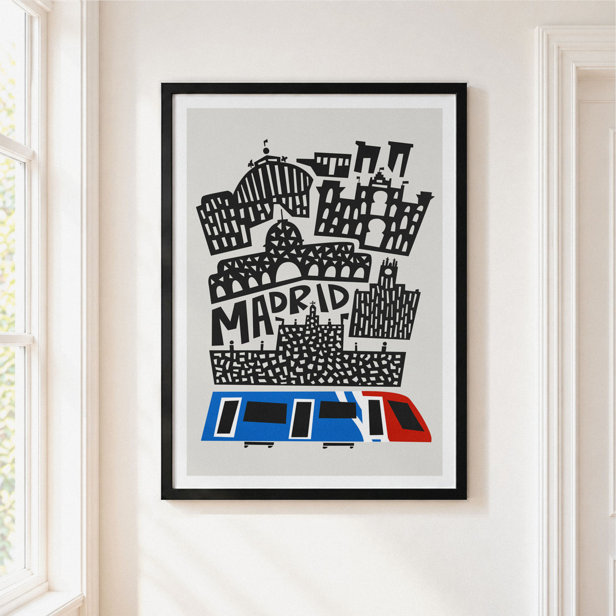 A framed art print featuring a stylized black and white illustration of Madrid landmarks with a blue and red tram at the bottom, hanging on a light beige wall next to a window.