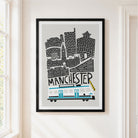A framed art print featuring a stylized Manchester skyline with a white and grey tram and black architectural details on a light gray background, hung on a cream-colored wall next to a window.
