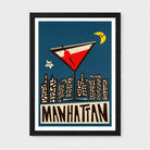 A framed art print featuring a stylized illustration of a martini glass above a Manhattan skyline, with a dark blue background, cream buildings, brown lettering reading "MANHATTAN," and a black frame on a light gray wall.