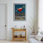 A framed vintage-style travel poster reading "MANHATTAN" hangs on a white wall above a gold bar cart in a bright living room. The poster features a dark blue background with a red and white airplane, black city skyline silhouettes, and a yellow crescent moon.