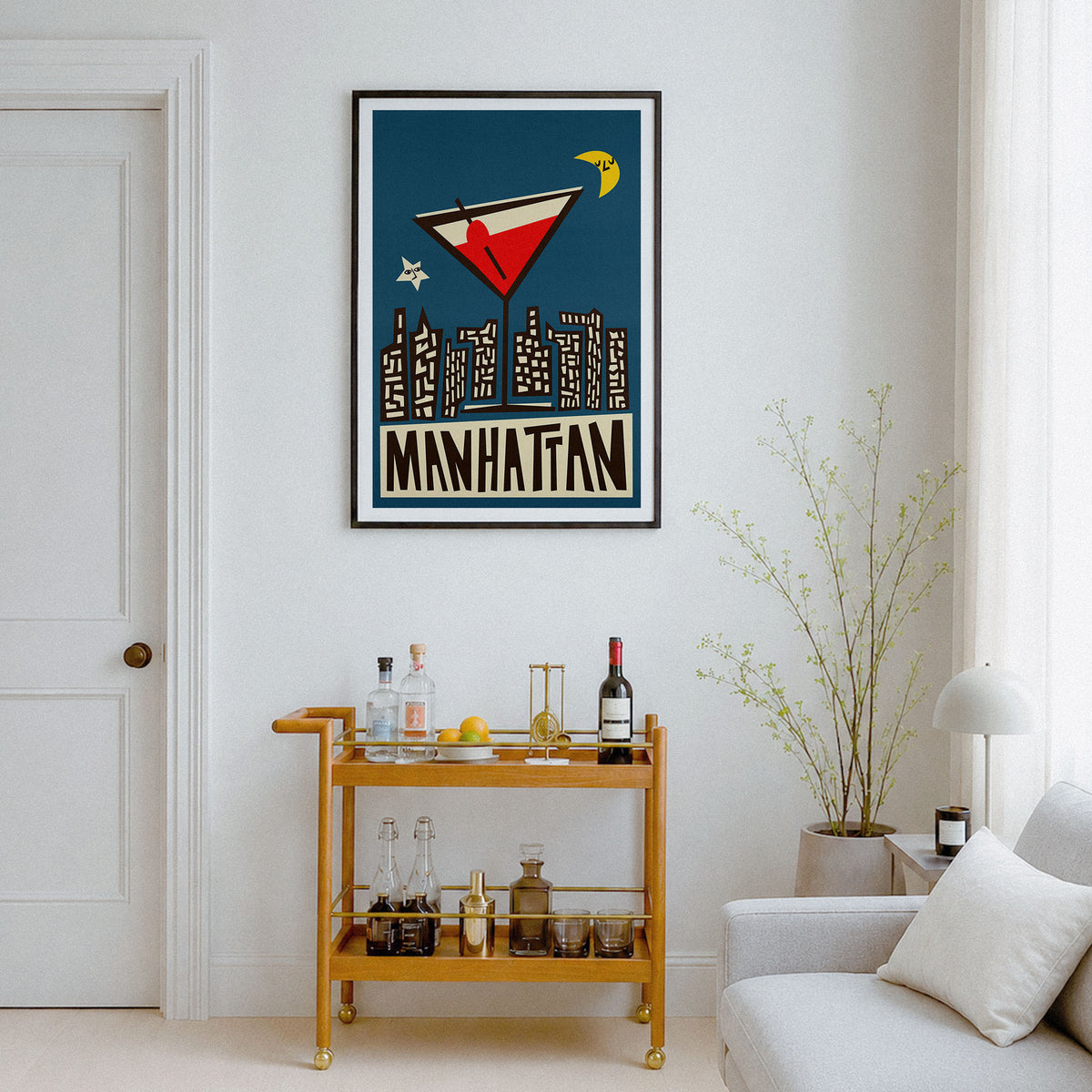 A framed vintage-style travel poster reading "MANHATTAN" hangs on a white wall above a gold bar cart in a bright living room. The poster features a dark blue background with a red and white airplane, black city skyline silhouettes, and a yellow crescent moon.