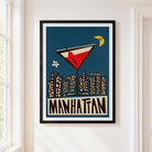 A framed art print featuring a retro-style Manhattan cocktail and city skyline on a dark blue background, hung on a white wall next to a window.