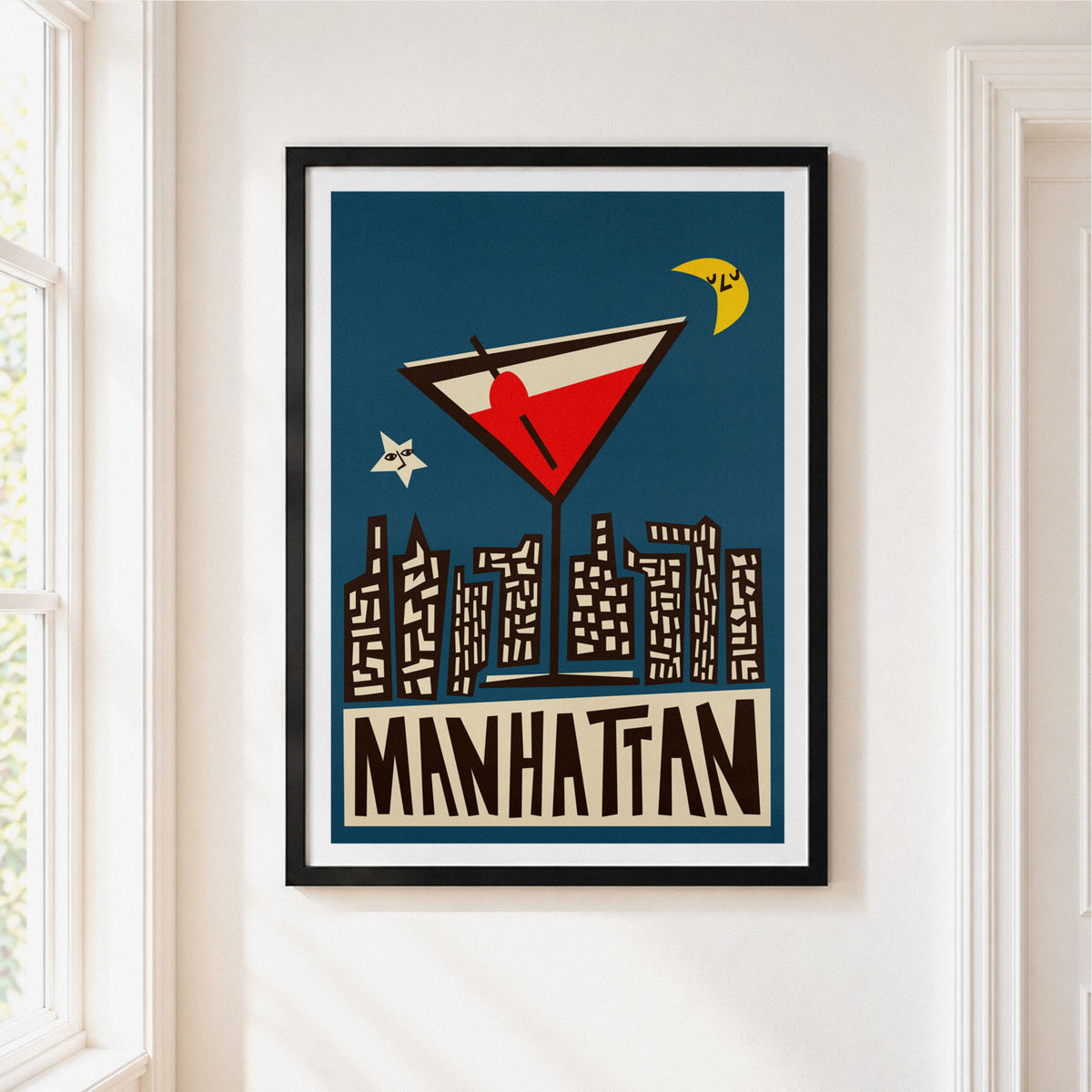 A framed art print featuring a retro-style Manhattan cocktail and city skyline on a dark blue background, hung on a white wall next to a window.