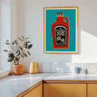 A framed art print featuring a stylized red hot sauce bottle on a teal background, hanging on a white kitchen wall above a countertop with a potted plant and mugs.