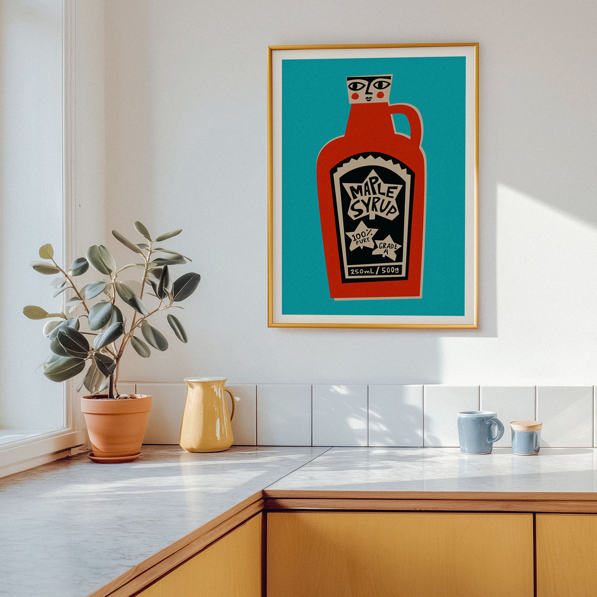 A framed art print featuring a stylized red hot sauce bottle on a teal background, hanging on a white kitchen wall above a countertop with a potted plant and mugs.
