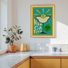 A framed art print featuring a colorful Margarita illustration hangs on a white kitchen wall above a counter with a potted plant and mugs.