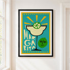 A framed art print featuring a retro-style Margarita cocktail illustration with a lime wheel and the word 'MARGARITA' on a turquoise background, hung on a white wall next to a window.