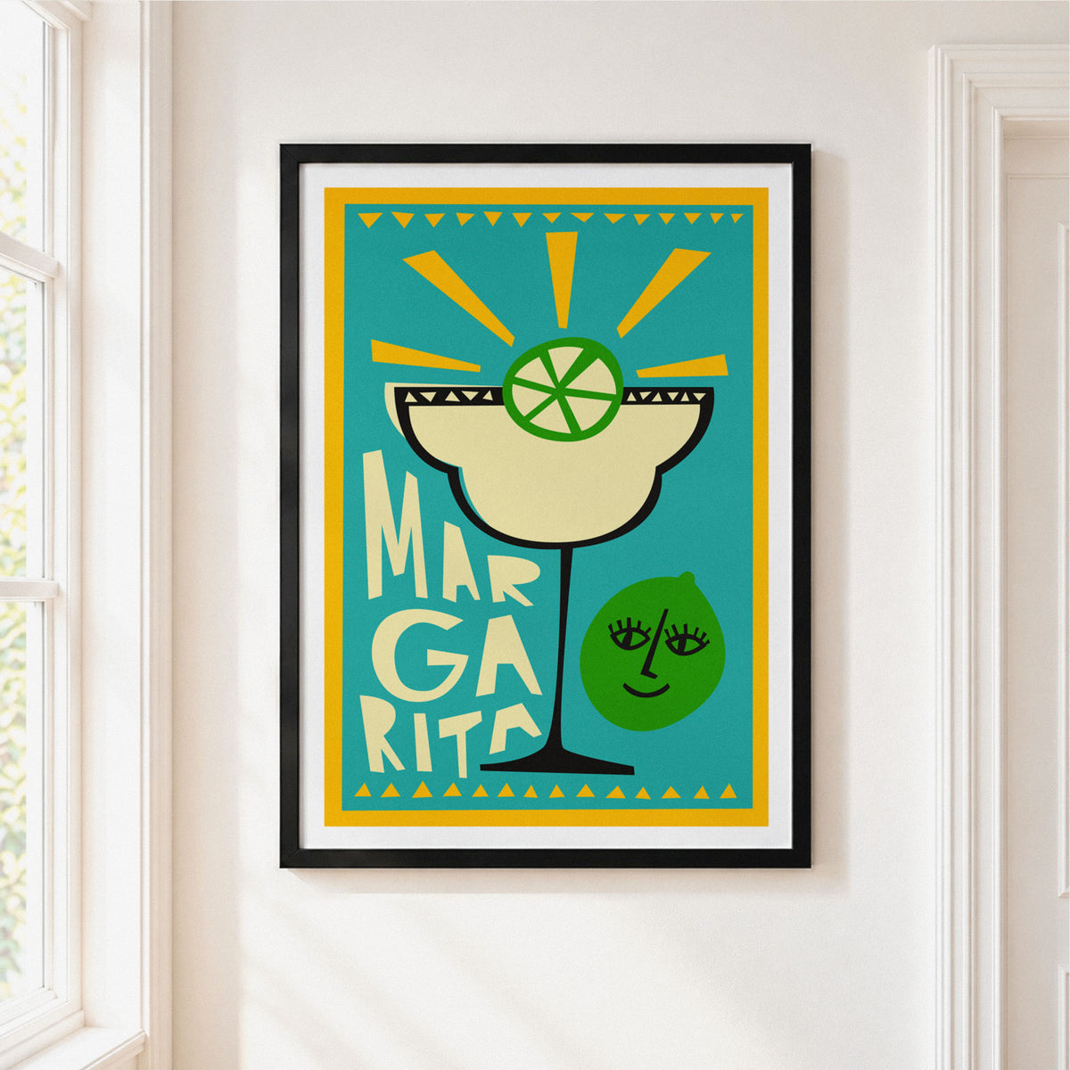 A framed art print featuring a retro-style Margarita cocktail illustration with a lime wheel and the word 'MARGARITA' on a turquoise background, hung on a white wall next to a window.