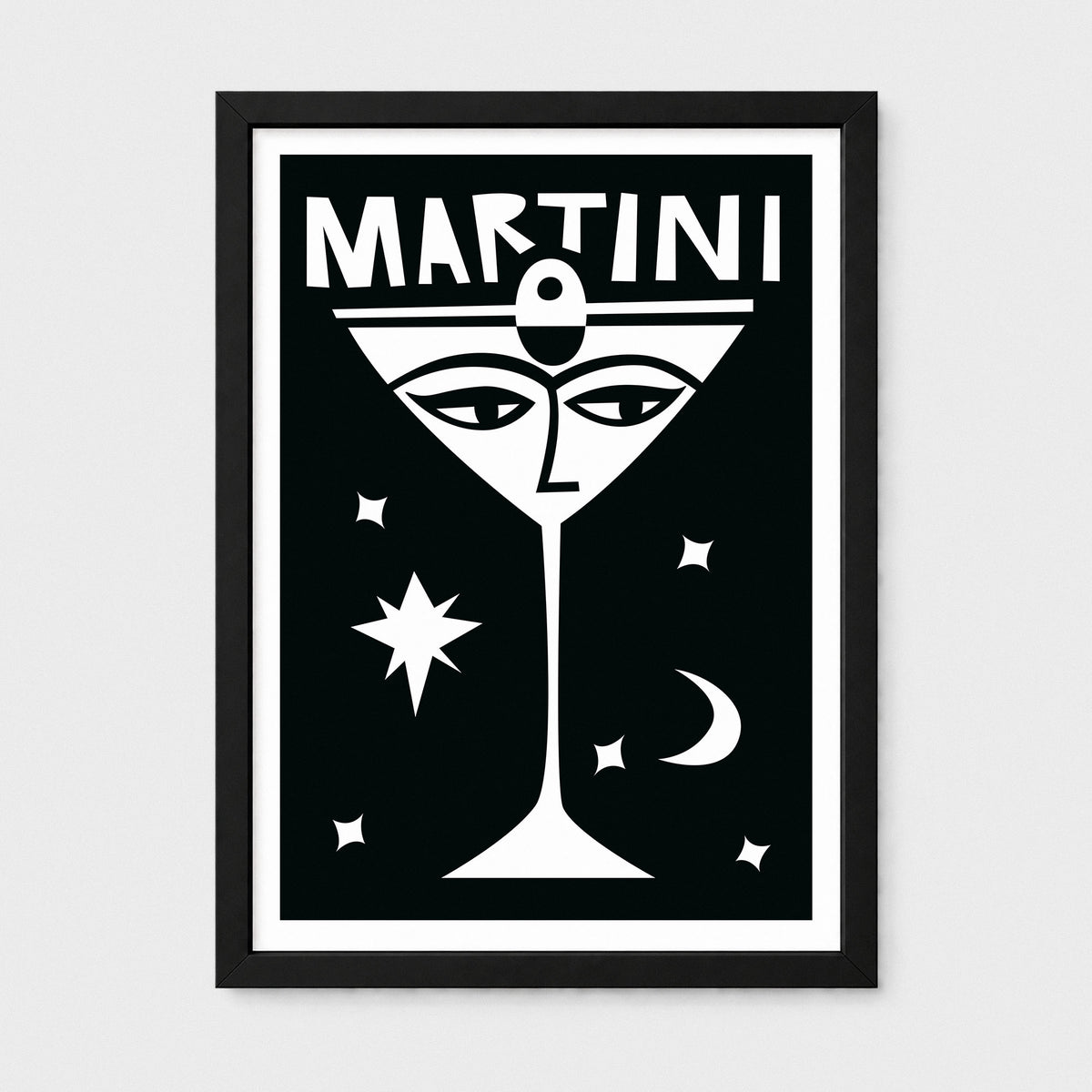 A framed black and white art print featuring a stylized martini glass with eyes, the word 'MARTINI' at the top, and small celestial shapes on a light gray background.