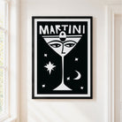 A framed black and white art print featuring a stylized martini glass with a face and the word 'MARTINI' at the top, hanging on a white wall next to a window.